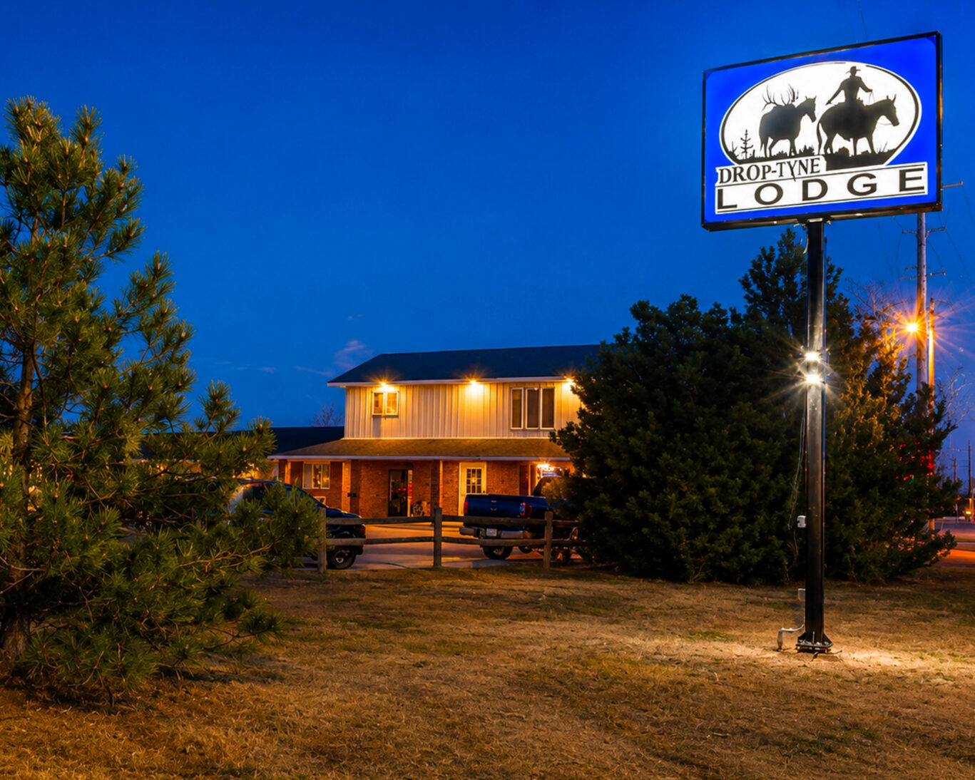 Evening exterior view of Drop-Tyne Lodge showing the lit sign, two-story building, and pine landscaping.