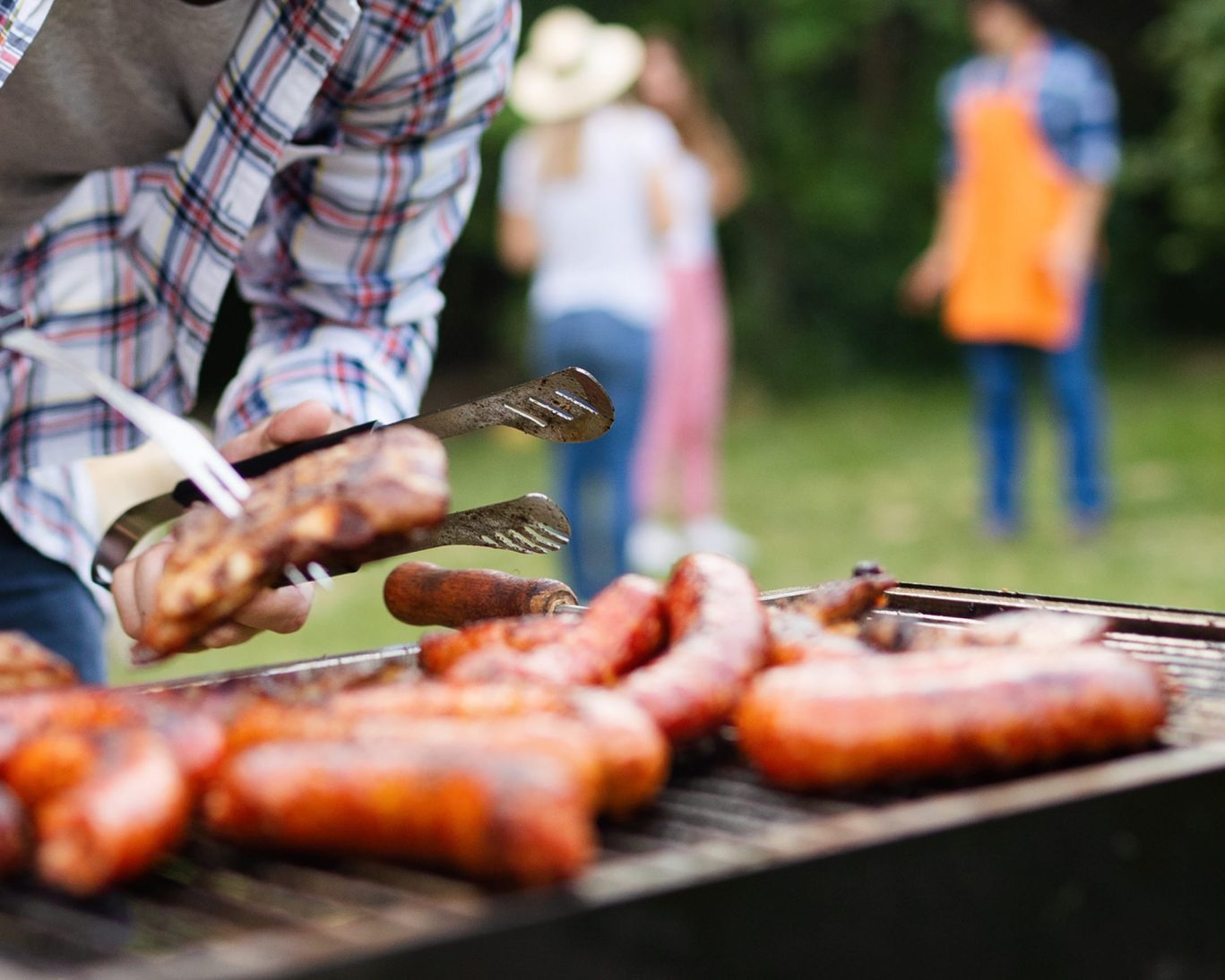 Close-up of sausages and meat grilling at outdoor barbecue with guests in the background at Huggy Bear Motel.
