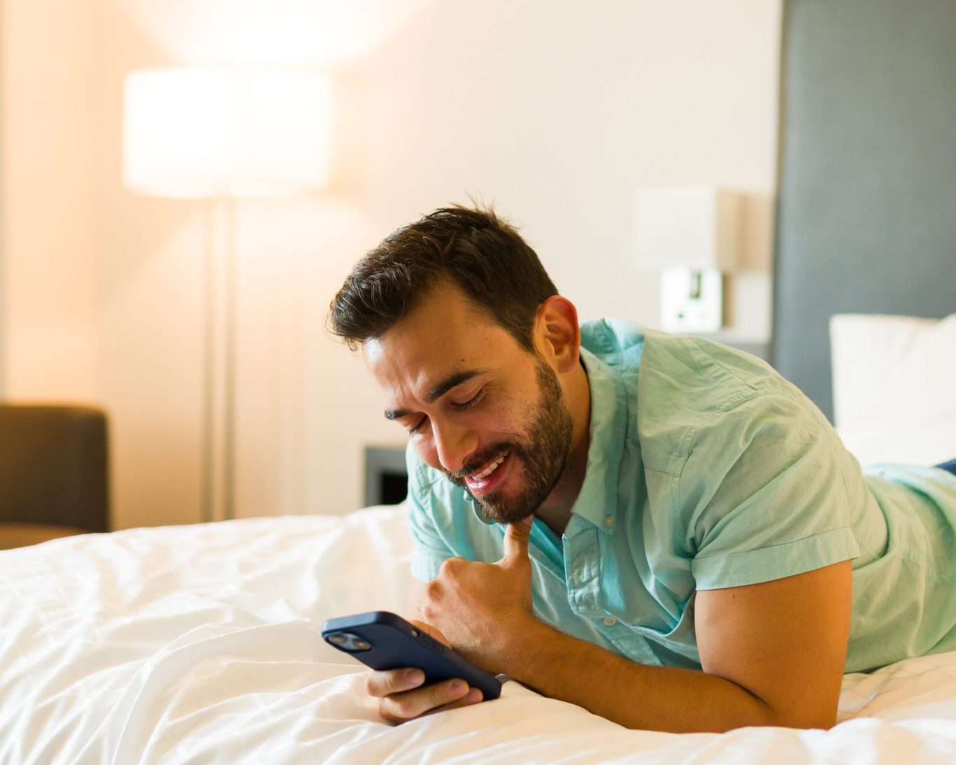 Man smiling and using smartphone while lying on hotel bed at Hampton motel.