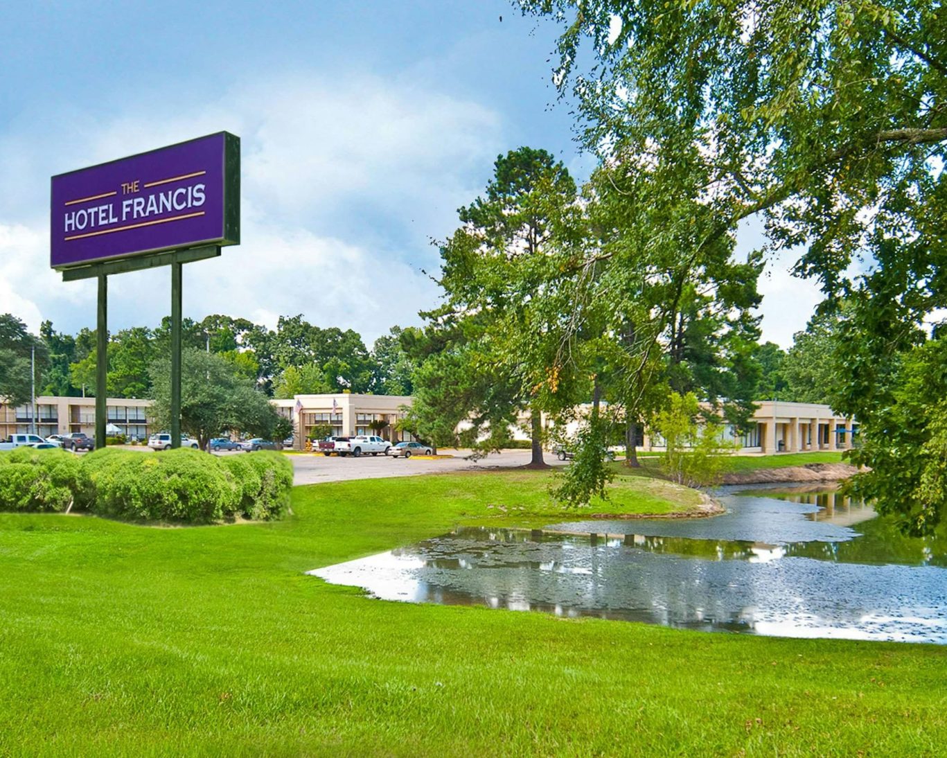Exterior view of The Hotel Francis with lush greenery, pond, and prominent roadside sign in Tatum, Texas.