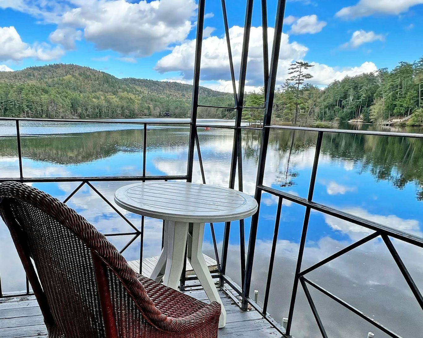 Lakefront balcony view with table and chair overlooking tranquil waters and forested hills.
