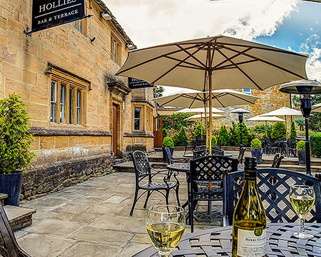 Patio seating at The Hollies Bar & Terrace with wine glasses and bottle set against a historic backdrop.
