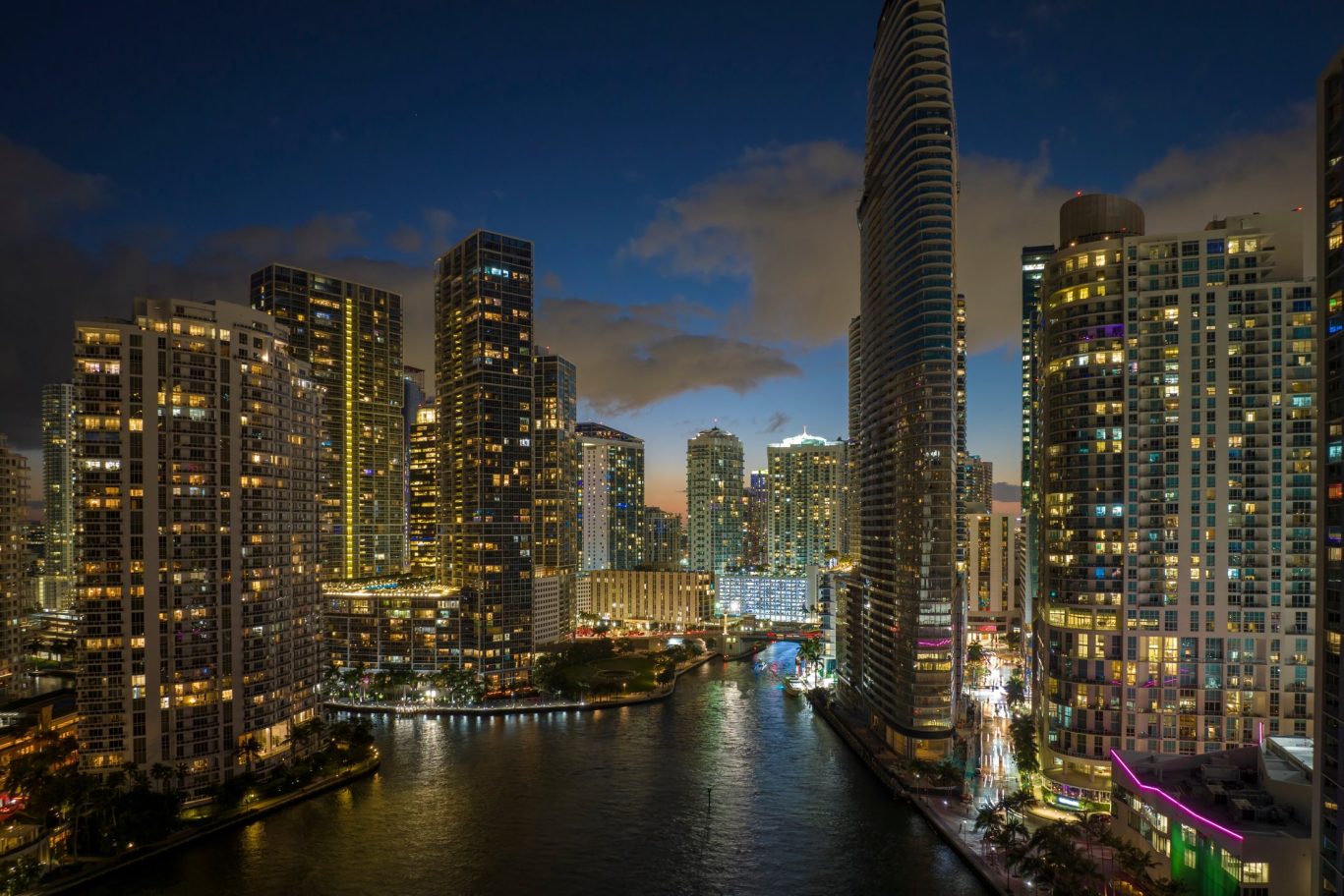 View from above of brightly illuminated skyscrapers in downtown Miami Brickell, Florida at night, with lights reflecting on the water.