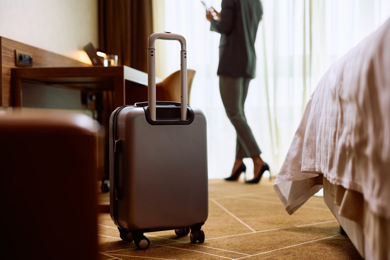 Rolling suitcase in upscale hotel room with businesswoman using phone by window.